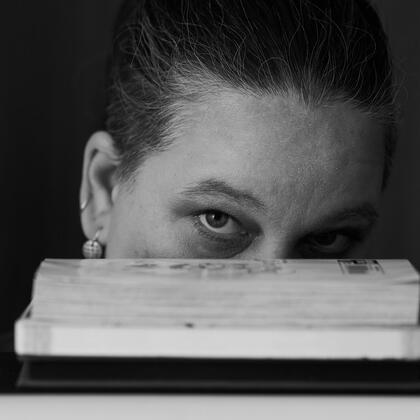 black and white image of a white-skinned woman peering out over a stack of books, nose and lower face obscured by books.